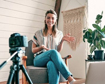 Influencer sind durch ihre Reichweite in der passenden Zielgruppe wichtige Markenbotschafter Eine lächelnde Frau sitzt in einem Sessel, während sie mit einer Kamera spricht. Vor ihr steht ein Laptop, und im Hintergrund sind Pflanzen und eine Wanddekoration sichtbar.