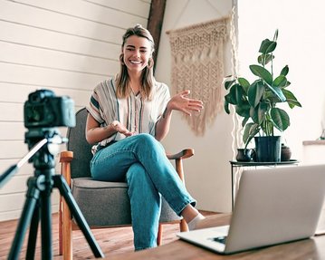 Medien im Wandel: Jeder kann heute Inhalte verbreiten Eine lächelnde Frau sitzt in einem Sessel, während sie mit einer Kamera spricht. Vor ihr steht ein Laptop, und im Hintergrund sind Pflanzen und eine Wanddekoration sichtbar.