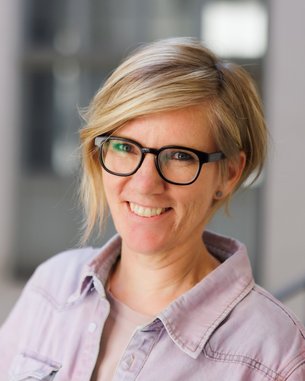 Eine lächelnde Frau mit blonden, schulterlangen Haaren und Brille trägt ein helles Hemd. Der Hintergrund ist unscharf und zeigt einen modernen Innenraum.