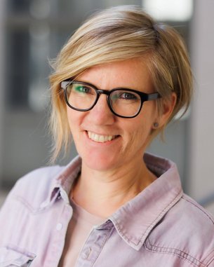 Eine lächelnde Frau mit blonden, schulterlangen Haaren und Brille trägt ein helles Hemd. Der Hintergrund ist unscharf und zeigt einen modernen Innenraum.