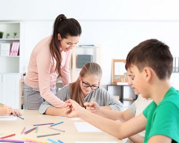 Eine Lehrerin unterstützt Schüler beim Zeichnen an einem Tisch, während sie verschiedene Buntstifte verwenden. Sechs Kinder konzentrieren sich auf ihre Arbeiten.
