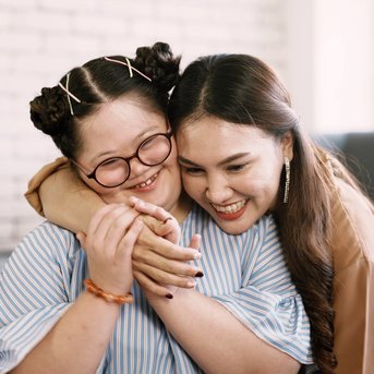 Zwei Frauen sitzen auf einem Sofa und umarmen sich lächelnd. Eine Frau trägt eine gestreifte Bluse und hat Zöpfe, während die andere eine braune Bluse trägt. Beide wirken freundlich und verbunden.