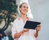Eine lächelnde Frau mit Brille hält ein schwarzes Tablet in der Hand, trägt ein helles Hemd und steht in einem modernen, hellen Raum. Im Hintergrund sind verschwommene Silhouetten von Personen zu sehen.