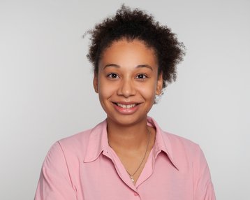 Junge Frau mit lockigem, braunem Haar und einem freundlichen Lächeln, trägt ein rosa Hemd. Der Hintergrund ist neutral und hell.