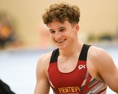 Ein junger Turner mit lockigem Haar trägt ein rotes Trikot mit dem Schriftzug "REIMER". Er lächelt und scheint sich über etwas zu freuen, während er in einer Turnhalle steht.