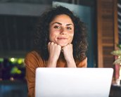 Eine Frau mit lockigem Haar sitzt vor einem Laptop, lächelt und stützt ihr Kinn auf ihre Hände. Der Hintergrund zeigt eine entspannte Umgebung mit Pflanzen.