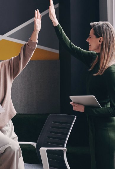 Zwei Frauen in einem modernen Büro geben sich ein High-Five. Eine Frau hält Dokumente, die andere ein Tablet. Farbige Wandgestaltung im Hintergrund.