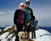 Ein Mann und eine Frau stehen auf einem schneebedeckten Berggipfel, beide sportlich gekleidet. Ein Golden Retriever sitzt vor ihnen. Im Hintergrund sind weitere Berglandschaften sichtbar.