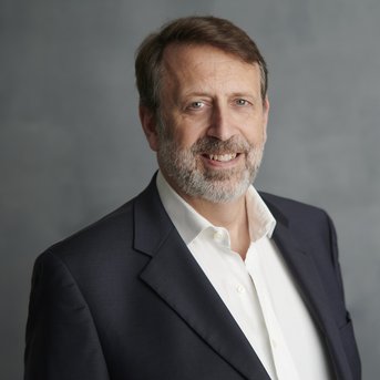 Ein Mann mit Bart trägt einen dunklen Anzug und ein weißes Hemd. Er steht vor einem neutralen Hintergrund und lächelt leicht.