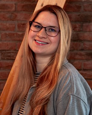 Eine lächelnde Frau mit langen, blonden Haaren und Brille steht vor einer Backsteinwand. Sie trägt ein graues Oberteil und hat eine gestreifte Bluse darunter.