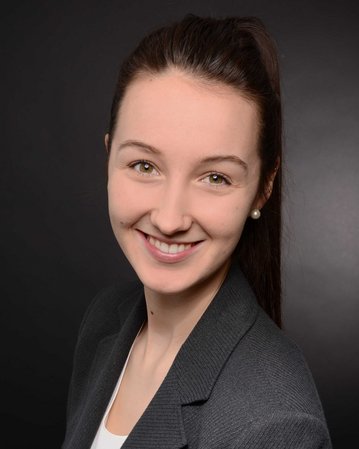 Junge Frau mit langen, braunen Haaren und Perlenohrringen, die in einem grauen Blazer lächelt, vor einem dunklen Hintergrund.