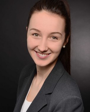 Junge Frau mit langen, braunen Haaren und Perlenohrringen, die in einem grauen Blazer lächelt, vor einem dunklen Hintergrund.