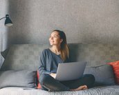 Eine junge Frau sitzt auf einem Sofa, lächelt und arbeitet an einem Laptop. Sie trägt einen grauen Pullover und hat ihre Beine angezogen. Im Hintergrund ist eine Lampe und ein dekorativer Vorhang zu sehen.
