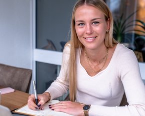 Eine junge Frau sitzt an einem Tisch, schreibt in ein Notizbuch und schaut lächelnd in die Kamera. Ein Laptop und eine Pflanze sind sichtbar.
