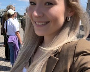Eine lächelnde Frau mit langen, blonden Haaren trägt ein braunes Blazer und einen Button mit der Aufschrift "HE PRINZIP". Im Hintergrund sind mehrere Personen und ein blauer Himmel zu sehen.