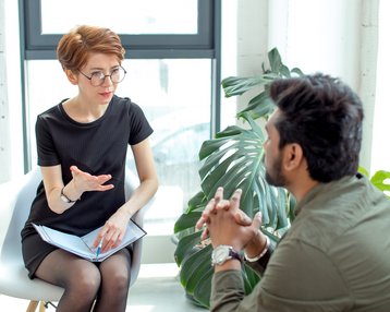 Eine Frau mit kurzen, roten Haaren und Brille sitzt auf einem Stuhl und spricht mit einem Mann, der ihr gegenüber sitzt. Beide scheinen in ein Gespräch vertieft zu sein.