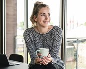 Eine Frau mit einem lächelnden Gesicht sitzt an einem Tisch und hält eine Tasse in der Hand. Sie trägt ein gemustertes Oberteil und hat ihre Haare locker frisiert.