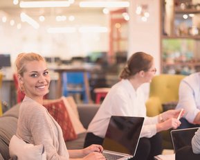 Fernstudium Master Medien- und Kommunikationsmanagement | SRH Fernhochschule Eine blonde Frau sitzt an einem Tisch in einem modernen Büro und lächelt, während sie an einem Laptop arbeitet. Im Hintergrund sind weitere Personen bei einer Besprechung zu sehen.