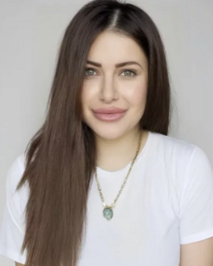Eine Frau mit langen, glatten Haaren trägt ein weißes T-Shirt und eine Halskette mit einem Anhänger. Sie lächelt in die Kamera.