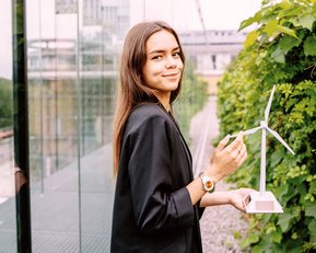 Eine junge Frau hält ein Modell einer Windturbine in der Hand und lächelt. Sie trägt eine schwarze Jacke und steht vor einer Glasfassade, umgeben von Pflanzen.