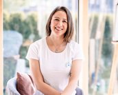 Eine Frau mit kurzen Haaren sitzt lächelnd auf einem Stuhl, trägt ein weißes T-Shirt und Jeans, umgeben von großen Fenstern.