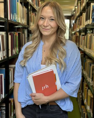 Eine lächelnde Frau mit langen, blonden Haaren steht in einer Bibliothek zwischen Bücherregalen und hält mehrere Notizbücher und einen Ordner in den Händen. Sie trägt ein gestreiftes Hemd und eine dunkle Hose.