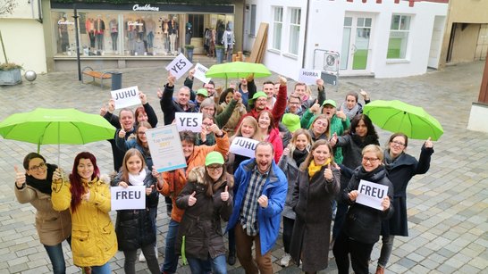 Eine Gruppe von Menschen hält grüne Regenschirme und Schilder mit den Aufschriften "JUHU", "YES" und "FREU" in einer Stadtstraße.