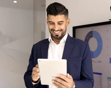 Ein Mann in einem Anzug hält ein Tablet und lächelt, während er vor einem Bildschirm mit Diagrammen und Grafiken steht.