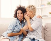 Zwei Frauen sitzen auf einem Sofa und lachen miteinander. Eine Frau hat lockige Haare und trägt eine Jeansjacke, während die andere kurze, blonde Haare hat und ein gestreiftes Hemd trägt.