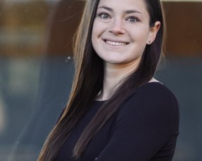 Eine Frau mit langen, dunklen Haaren lächelt freundlich, während sie mit verschränkten Armen in einem schwarzen Oberteil posiert. Der Hintergrund ist unscharf und zeigt eine helle, moderne Umgebung.