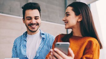 Ein Mann und eine Frau stehen nebeneinander, beide halten elektronische Geräte in der Hand. Der Mann lächelt, während die Frau auf ihr Smartphone schaut. Sie scheinen sich zu unterhalten.