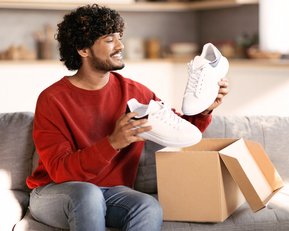Ein Mann sitzt auf einem Sofa und hält ein Paar weiße Schuhe in den Händen, während er eine geöffnete Kartonbox neben sich hat.