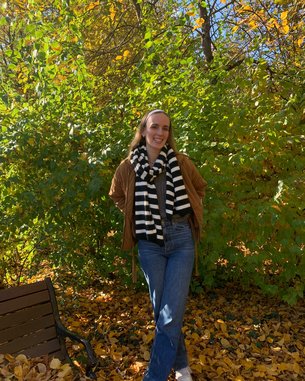 Eine Person steht in einem herbstlichen Park, trägt eine braune Jacke und einen schwarz-weißen gestreiften Schal. Im Hintergrund sind grüne Sträucher und gelbe Blätter zu sehen.