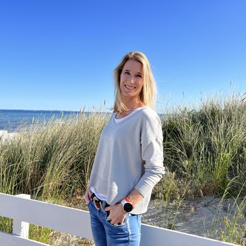 Eine Frau mit langen, blonden Haaren steht an einem Strand, umgeben von Gras und einem weißen Zaun. Sie trägt einen grauen Pullover und Jeans, während sie in die Kamera lächelt.