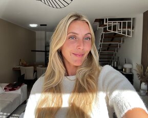 Eine Person mit langen, blonden Haaren lächelt in die Kamera, während sie in einem hellen Raum mit moderner Einrichtung steht.
