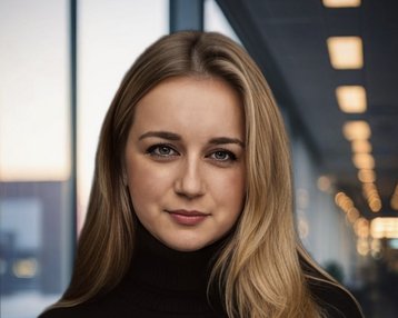 Eine junge Frau mit langen, blonden Haaren trägt einen schwarzen Rollkragenpullover. Sie blickt direkt in die Kamera, während unscharfe Lichter im Hintergrund sichtbar sind.