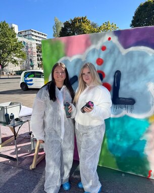 Zwei Frauen in weißen Schutzanzügen halten Spraydosen vor einer bemalten Wand. Im Hintergrund sind Bäume und ein Fahrzeug sichtbar.