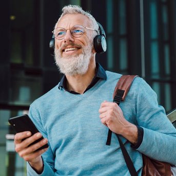 Ein älterer Mann mit Brille und Kopfhörern lächelt, während er ein Smartphone in der Hand hält. Er trägt einen hellblauen Pullover und eine braune Tasche.