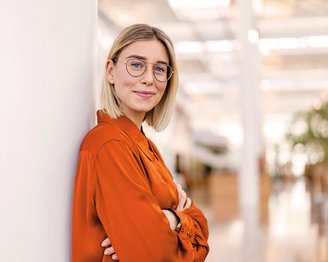 Eine junge Frau mit blonden, schulterlangen Haaren und Brille steht lächelnd an einer Wand. Sie trägt ein orangefarbenes Hemd und hat die Arme vor der Brust verschränkt.