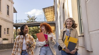 Drei Frauen gehen lächelnd auf einem Gehweg, tragen Ordner und einen Kaffeebecher, umgeben von Gebäuden und Tageslicht.