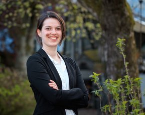 Eine Person mit kurzen Haaren trägt einen schwarzen Blazer und steht mit verschränkten Armen in einer grünen Umgebung. Sie lächelt freundlich in die Kamera.