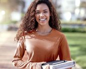 Eine junge Frau mit lockigem Haar trägt mehrere Bücher und Notizblöcke in den Händen. Sie lächelt und steht auf einem Gehweg, umgeben von einer grünen Umgebung.