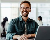 Ein lächelnder Mann mit Brille sitzt an einem Tisch, arbeitet an einem Laptop und hat ein Notizbuch vor sich. Im Hintergrund sind weitere Personen in einem modernen Büro zu sehen.