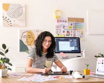 Eine Frau mit lockigem Haar sitzt an einem Tisch und arbeitet mit einem Grafiktablet. Um sie herum liegen verschiedene Designmaterialien und Farbmuster. Im Hintergrund sind Kunstwerke und ein Computerbildschirm sichtbar.