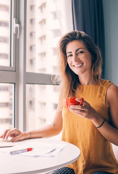 Eine Frau mit langen Haaren sitzt an einem Tisch, hält eine Tasse in der Hand und arbeitet an einem Laptop, während sie aus dem Fenster schaut.