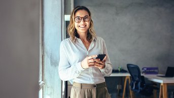 Eine Frau mit Brille steht lächelnd an einem Fenster und hält ein Smartphone in der Hand. Sie trägt ein gestreiftes Hemd und eine elegante Hose. Im Hintergrund sind Büromöbel sichtbar.