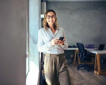 Eine Frau mit Brille steht lächelnd an einem Fenster und hält ein Smartphone in der Hand. Sie trägt ein gestreiftes Hemd und eine elegante Hose. Im Hintergrund sind Büromöbel sichtbar.