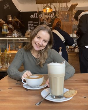 Eine lächelnde Frau sitzt an einem Tisch in einem Café, vor ihr stehen eine Tasse Kaffee und ein Glas Latte Macchiato, begleitet von einem Keks. Im Hintergrund sind weitere Café-Möbel und Gäste sichtbar.