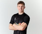 Ein junger Mann mit kurzem, lockigem Haar trägt ein schwarzes T-Shirt und steht mit verschränkten Armen. Er lächelt freundlich in die Kamera.
