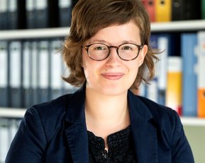 Eine Person mit kurzen, lockigen Haaren und Brille trägt einen dunklen Blazer. Im Hintergrund sind Aktenordner in verschiedenen Farben sichtbar.
